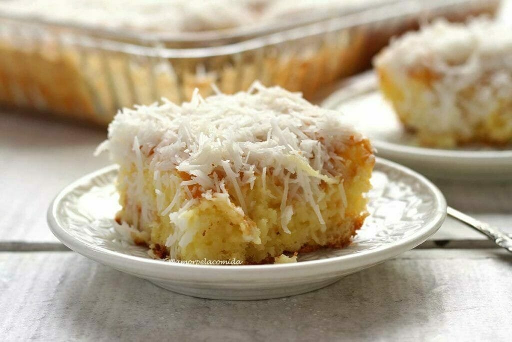 Pedaço de bolo de coco com cobertura de coco em prato redondo, ao fundo a assadeira de vidro e um pratinho com mais um pedaço de bolo desfocados