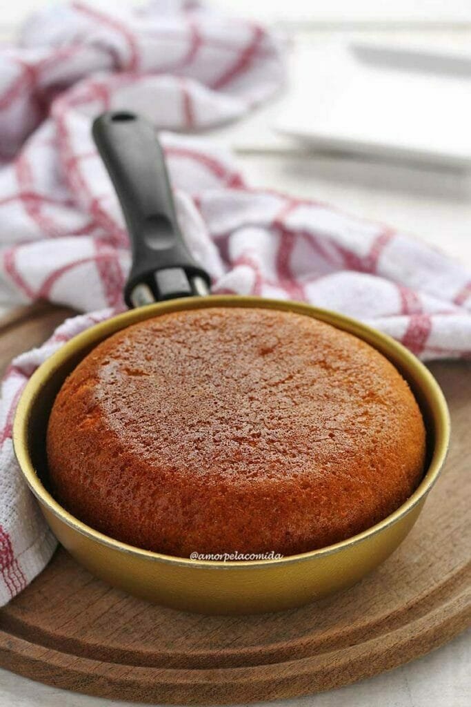 Frigideira dourada com cabo preto sobre tábua de madeira, dentro da frigideira um bolo de fubá com banana