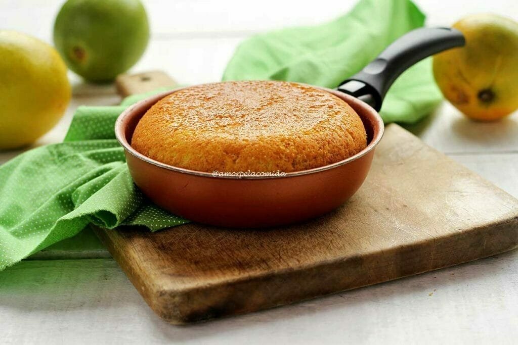 Frigideira marrom pequena com bolo de laranja dentro sobre uma tábua de madeira e um pano verde claro ao lado, algumas laranjas desfocadas ao fundo em uma mesa branca