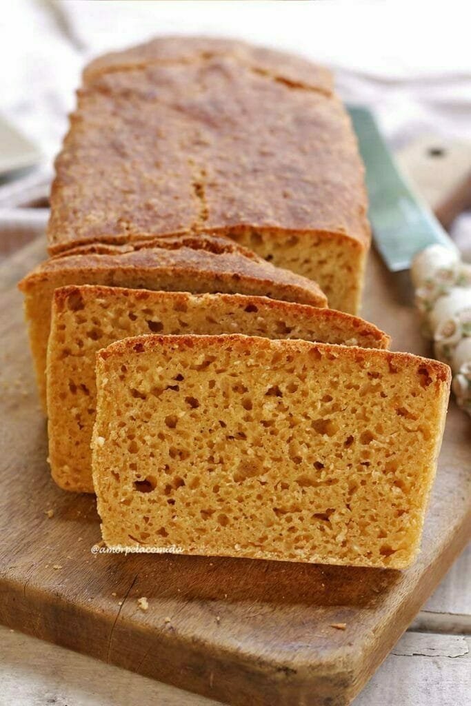 Pão fatiado sobre tábua de madeira em uma mesa branca, ao lado do pão uma faca com cabo branco decorado