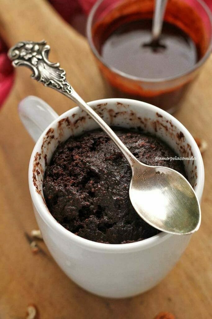 Xícara branca com bolo de chocolate dentro assado no micro-ondas, no topo da borda da xícara uma colher de prata antiga com o cabo decorado