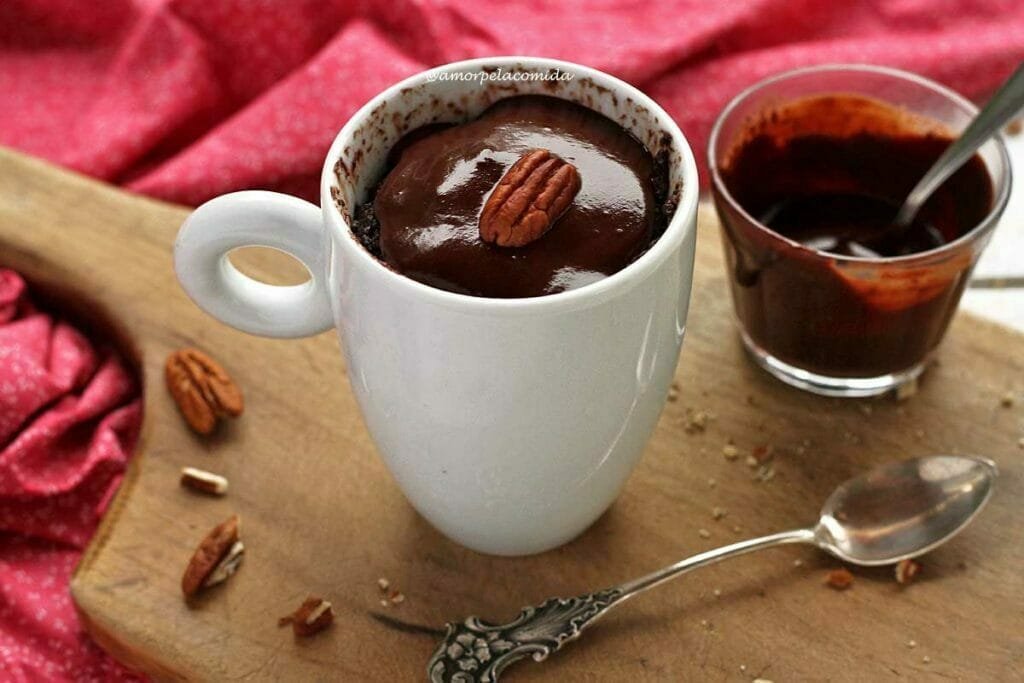 Xícara branca com bolo de chocolate dentro, o bolo leva cobertura de chocolate cremoso e tem uma noz pecan decorando no topo. A xícara está apoiada em uma tábua de madeira