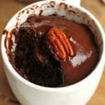 Xícara branca com bolo de chocolate dentro, no topo do bolo uma cobertura de chocolate e uma noz pecan, o bolo já está parcialmente comido
