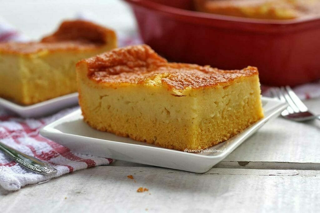 Pedaço de bolo de fubá cremoso sobre prato quadrado branco pequeno, ao fundo mais uma fatia e a forma vermelha com o restante do bolo dentro