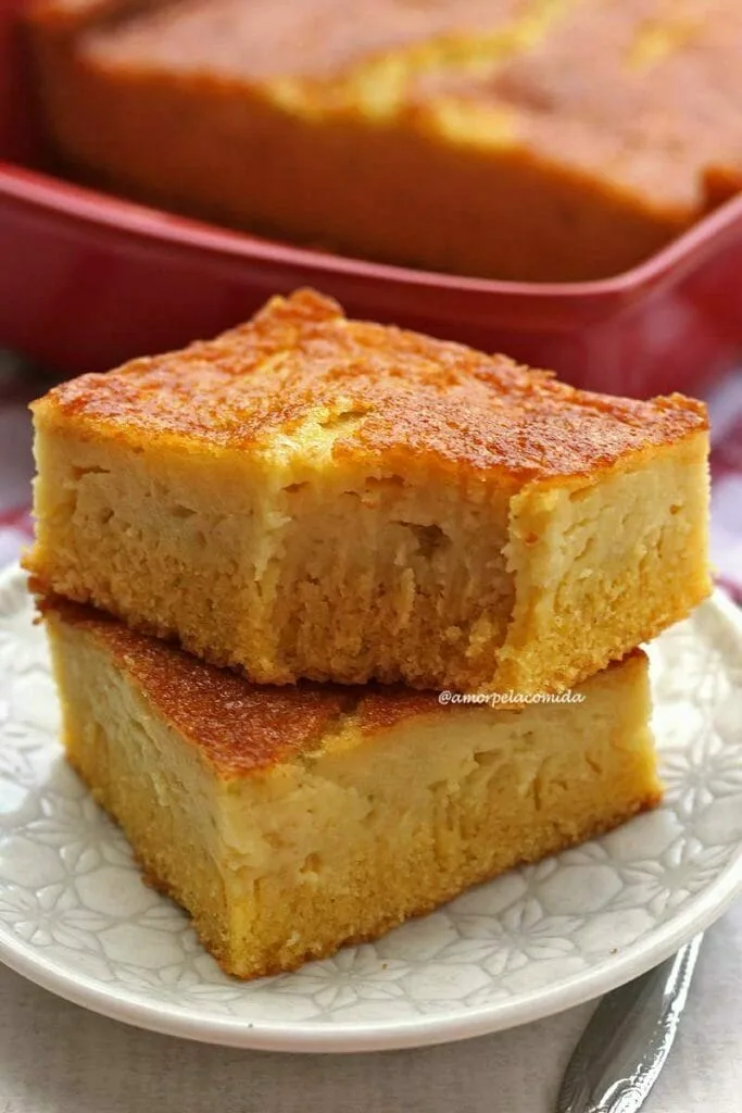 Duas fatias de bolo de fubá cremoso sobrepostas em um prato redondo pequeno, ao fundo a forma vermelha com o bolo dentro sobre uma mesa branca