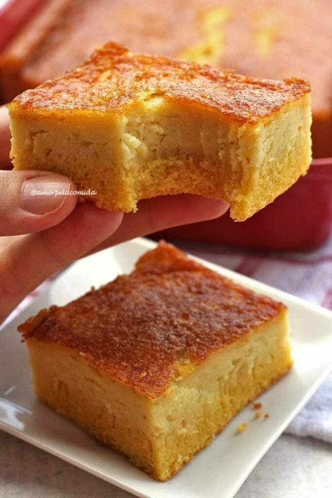 Mão segurando pedaço de bolo de fubá com laranja mordido, abaixo mais uma fatia sobre prato quadrado pequeno, ao fundo uma forma vermelha sobre uma mesa branca