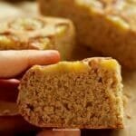 Mão segurando pedaço de bolo de banana cortado, no todo da fatia algumas rodelas de banana, ao fundo o restante do bolo