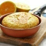 Frigideira marrom com cabo preto sobre tábua de madeira de cor clara, dentro bolo de laranja e ao fundo uma laranja partida ao meio