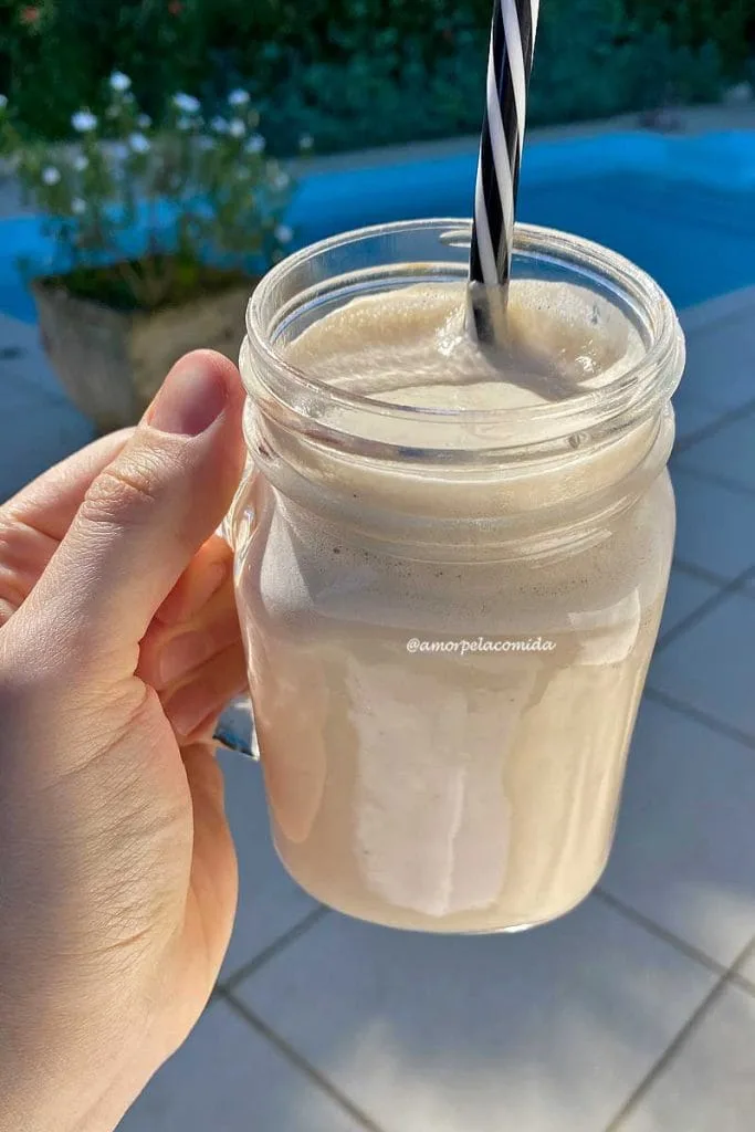 Mão segurando copo quadrado com canudo preto e branco dentro, cheio de leite vegetal de castanha