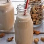 Garafinha de vidro com leite vegetal dentro com canudo vermelho e branco, ao fundo o pote de castanha de caju e mais um copo de leite