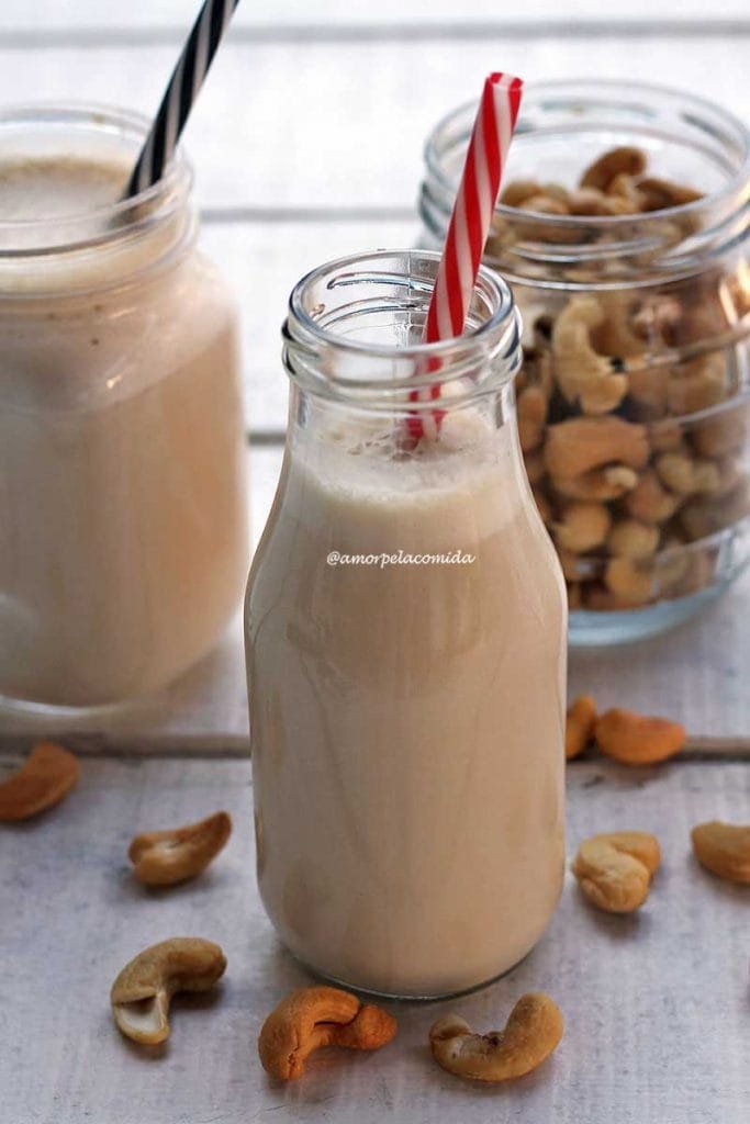 Garafinha de vidro com leite vegetal dentro com canudo vermelho e branco, ao fundo o pote de castanha de caju e mais um copo de leite