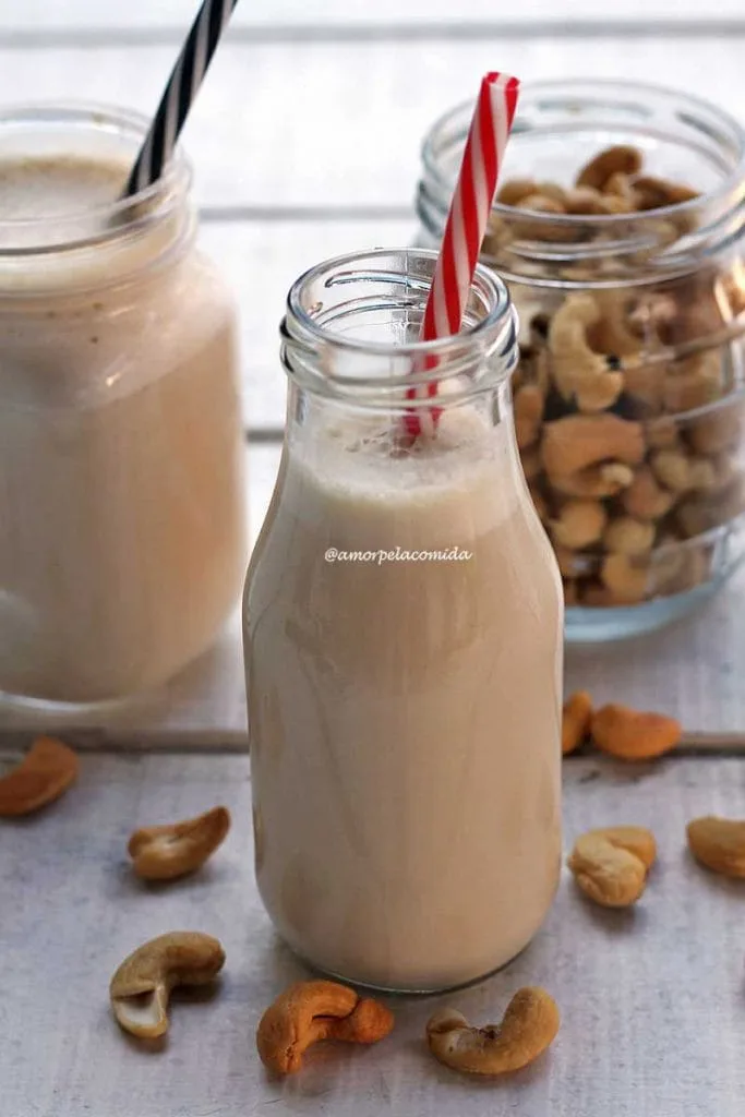 Pequeña botella de cristal con leche vegetal en su interior con pajita roja y blanca, al fondo el tarro de anacardos y otro vaso de leche