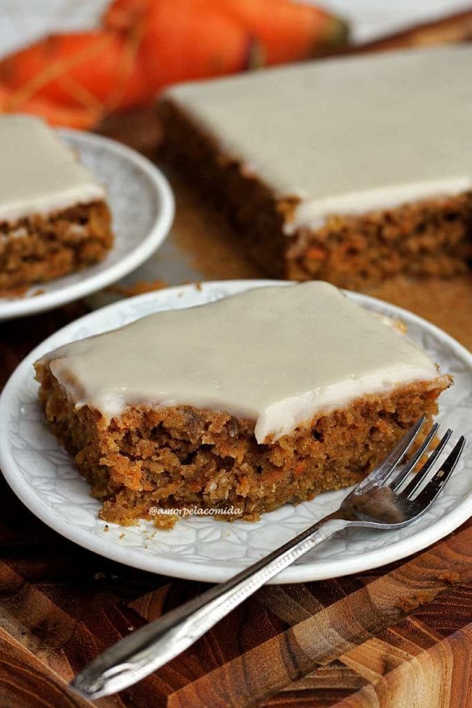 Pedaço de bolo de cenoura com textura rústica e cobertura branca que é cítrica. O bolo está em um prato redondo branco e pequeno ao fundo mais um pedaço de bolo em um prato e o restante do bolo em uma tábua