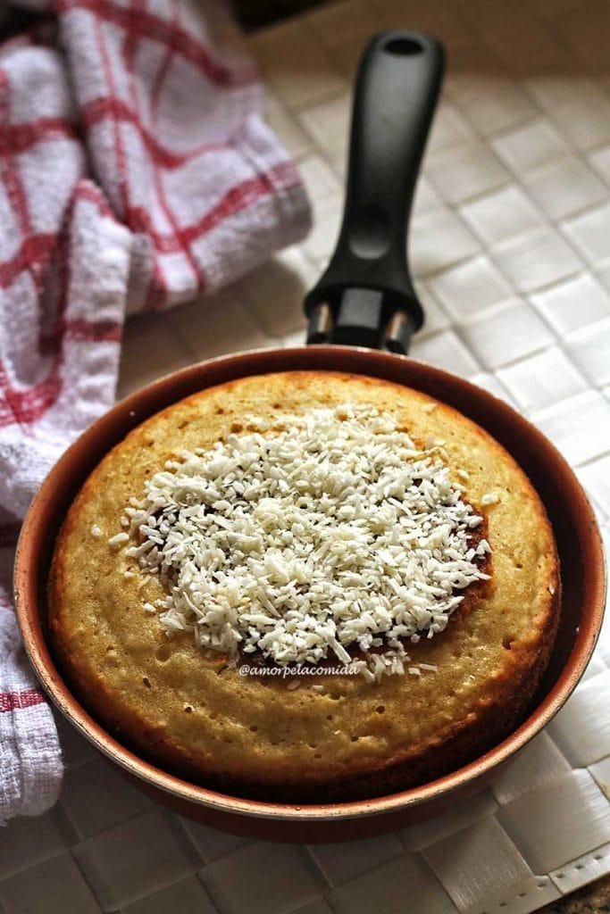 Frigideira pequena com bolo de coco dentro, sobre o bolo flocos de coco, abaixo da frigideira um jogo americano branco quadriculado e uma toalha branca com xadrez vermelho