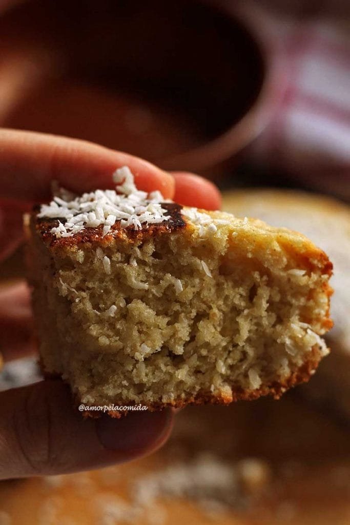 Mão segurando pedaço triangular pequeno de bolo de coco, no topo do bolo alguns flocos de coco