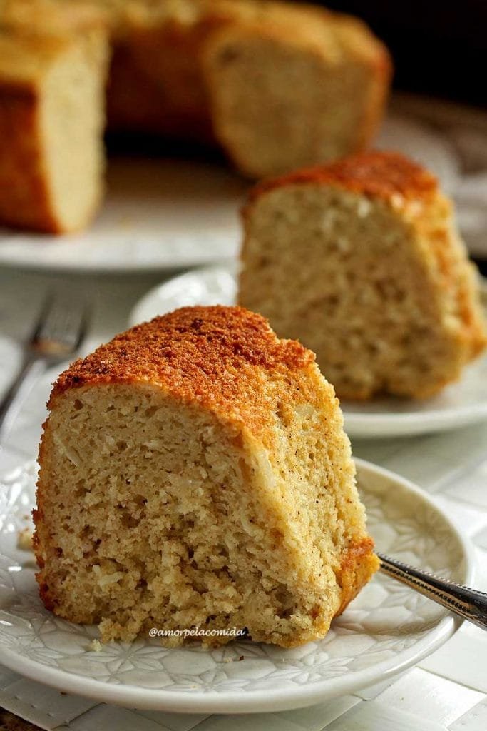 Duas fatias de bolo de banana e coco sobre dois pratinhos redondos pequenos, ao fundo o bolo partido desfocado