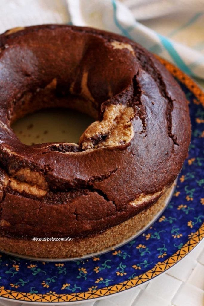 Bolo redondo com furo no meio, na parte superior é mesclado com massa de chocolate, o bolo está sobre um prato branco com borda azul e detalhes amarelos