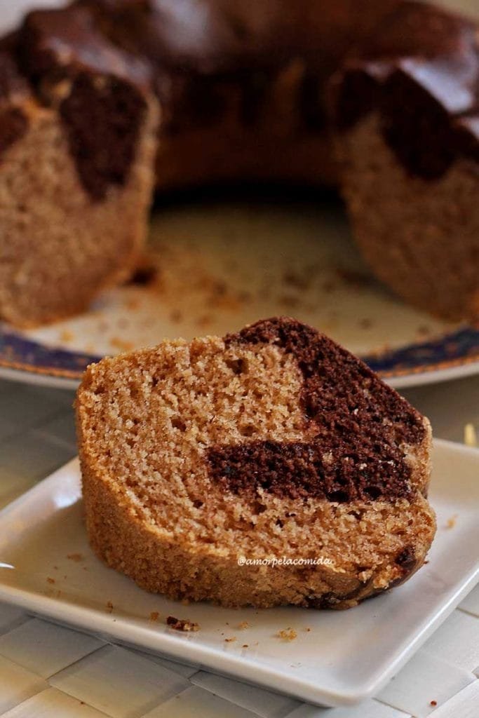 Fatia de bolo de canela com parte mesclada de chocolate sobre prato branco quadrado pequeno, ao fundo o bolo partido