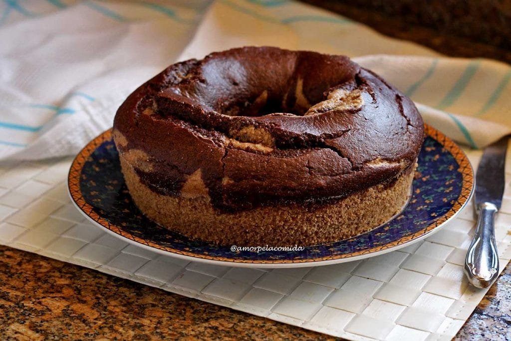 Bolo redondo com furo no meio, na parte superior é mesclado com massa de chocolate, o bolo está sobre um prato branco com borda azul e detalhes amarelos sobre jogo americano branco em uma bancada de mármore marrom