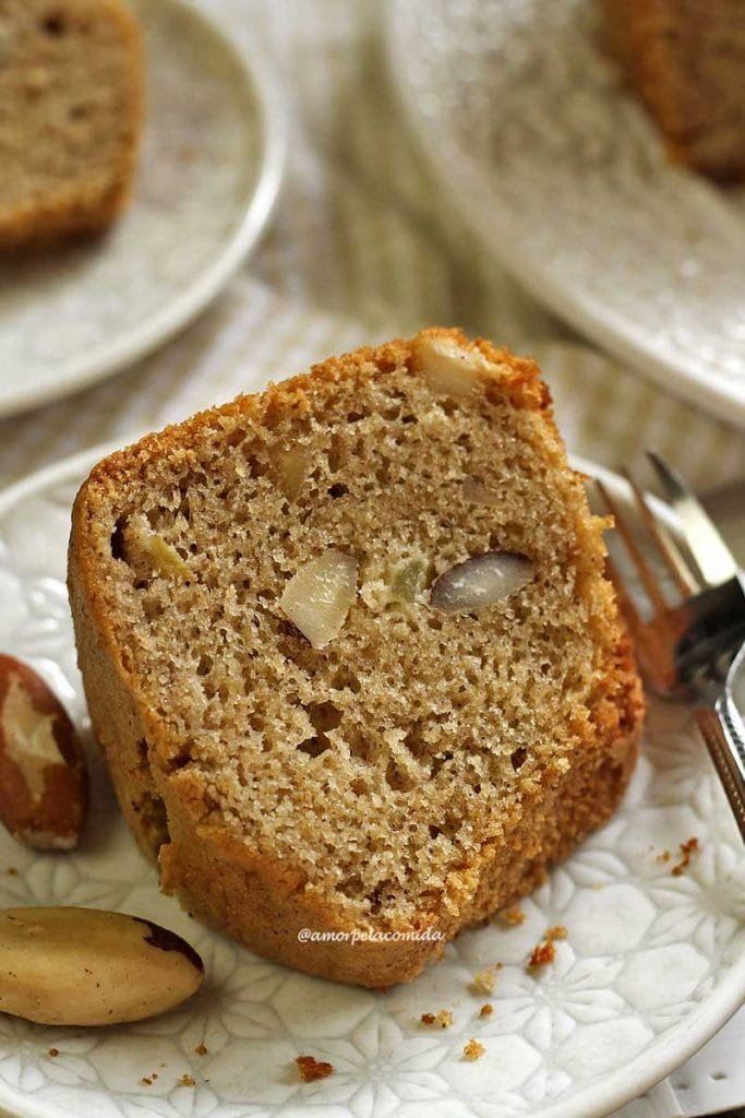 Pedaço de bolo de castanha sobre prato branco redondo pequeno com um garfinho ao lado e algumas castanhas