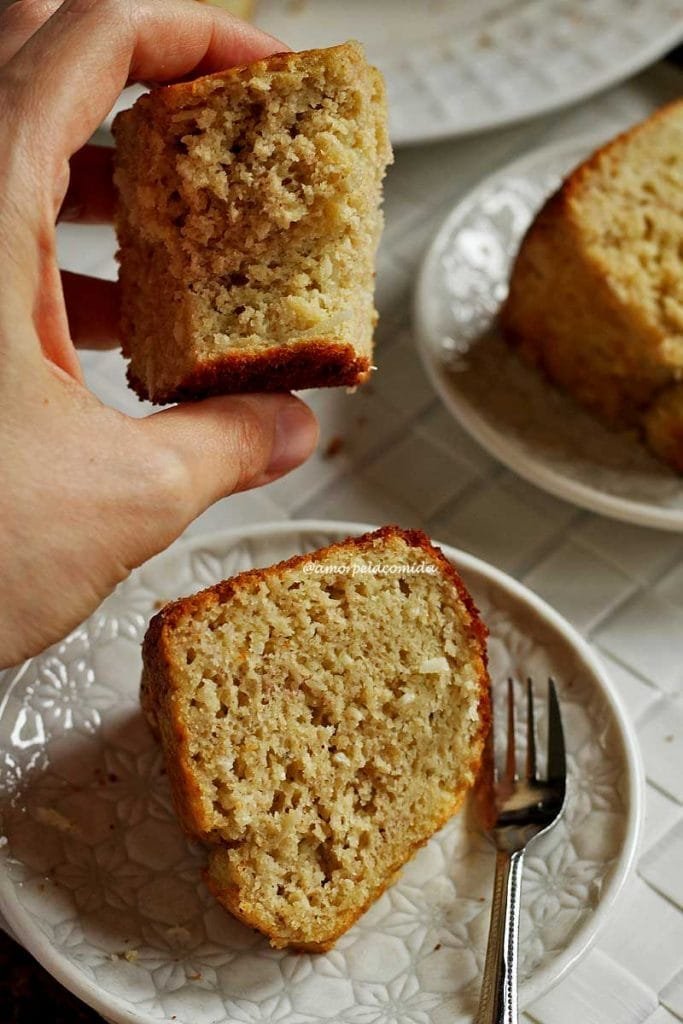 Mão segurando pedaço mordido de bolo de banana com coco, abaixo duas fatias de bolo deitadas sobre dois pratos brancos pequenos redondos