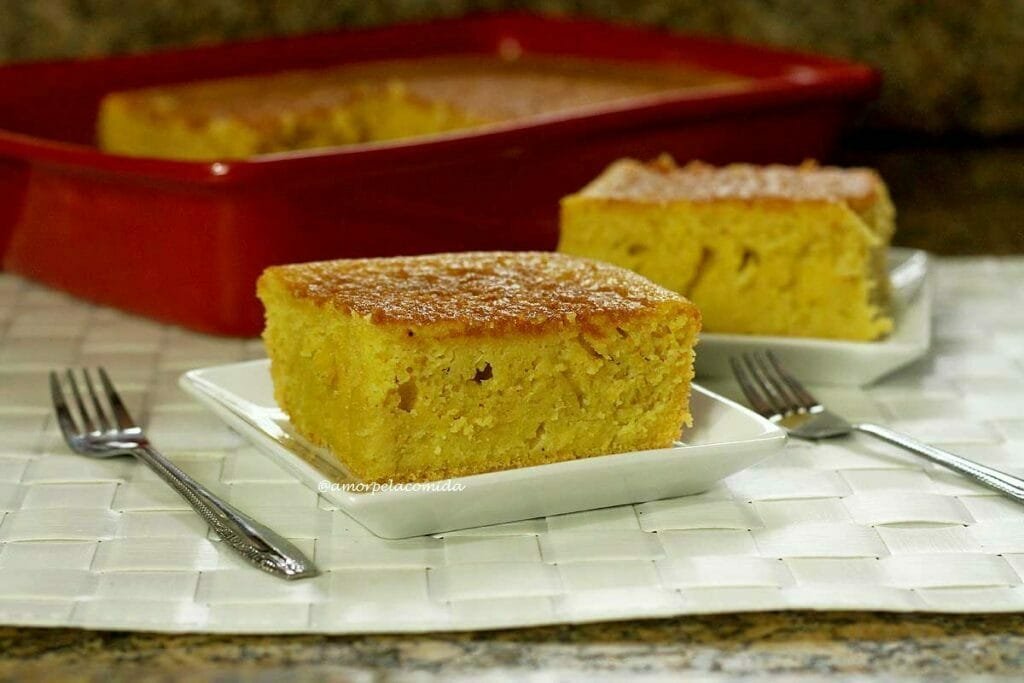 Dois pedaços de bolo de pamonha o sobre prato quadrado pequeno, ao fundo a forma vermelha com o restante do bolo