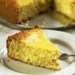 Fatia de bolo de queijo sobre prato branco redondo, ao fundo o bolo partido