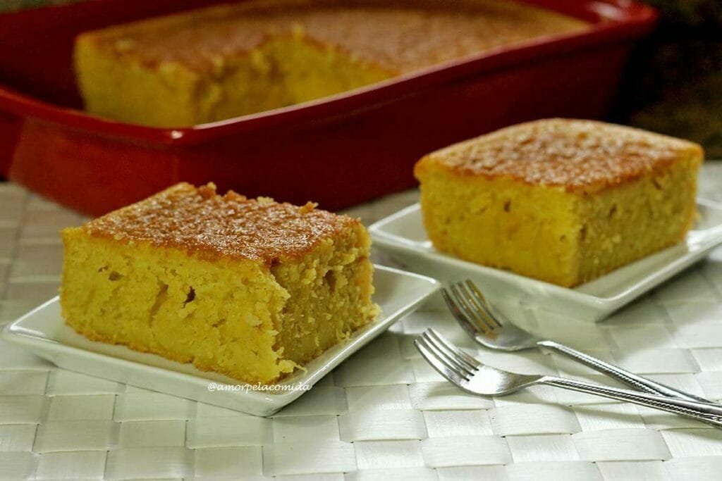 Dois pedaços de bolo de pamonha o sobre prato quadrado pequeno, ao fundo a forma vermelha com o restante do bolo