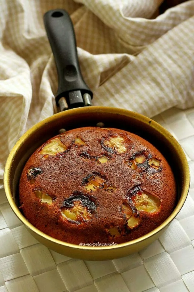 Frigideira dourada sobre jogo americano branco, dentro da frigideira um bolo de banana e maçã, no topo do bolo pedaços de maçã e rodelas de banana