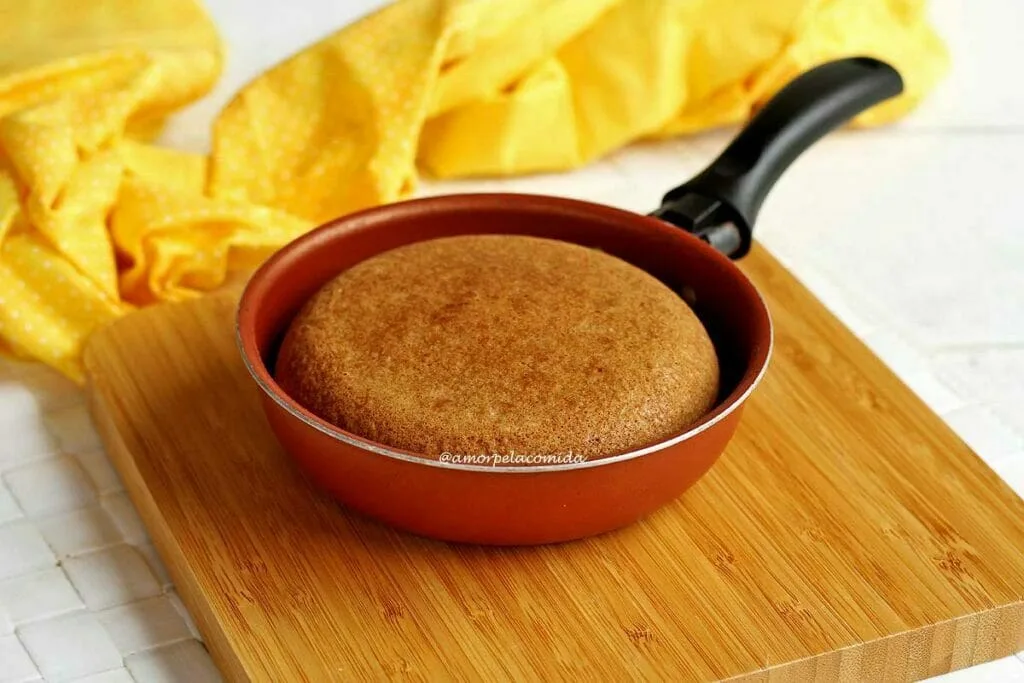 Frigideira pequena com bolo de fubá dentro sobre tábua quadrada de madeira em uma mesa branca, ao fundo um pano amarelo dobrado