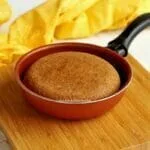 Frigideira pequena com bolo de fubá dentro sobre tábua quadrada de madeira em uma mesa branca, ao fundo um pano amarelo dobrado