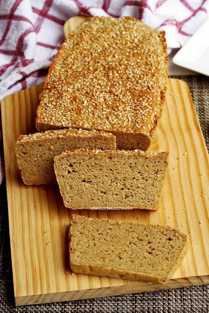 Três fatias de pão apoiadas sobre o pão que no topo tem gergelim e está sobre uma tábua de madeira