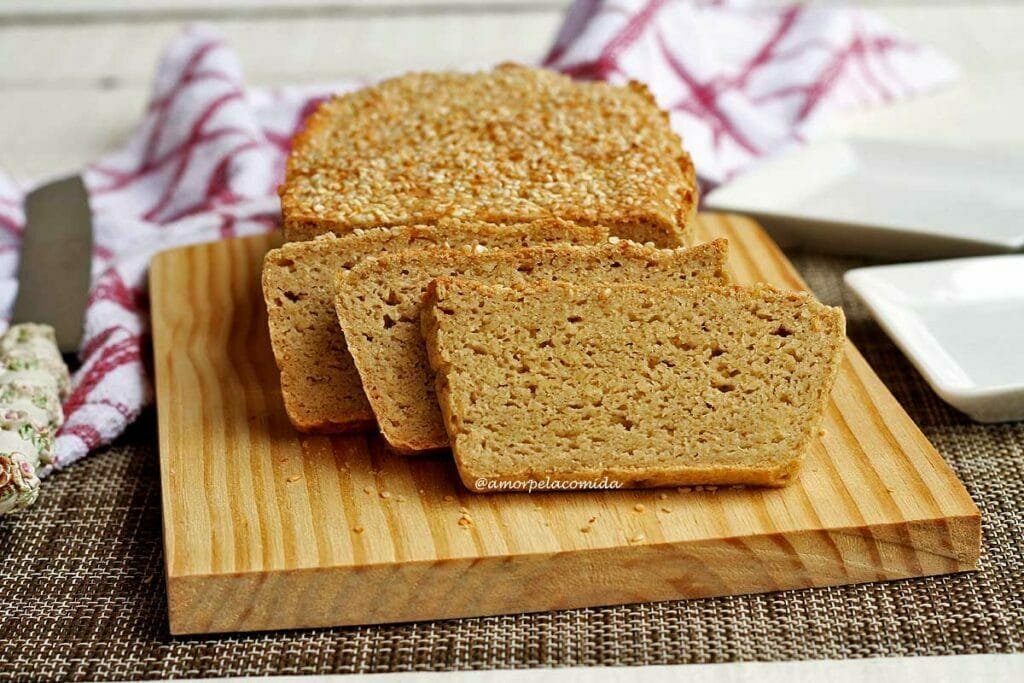 Pão com três fatias apoiadas sobre tábua de madeira, abaixo da tábua um jogo americano marrom, ao lado dois pratos quadrados pequenos