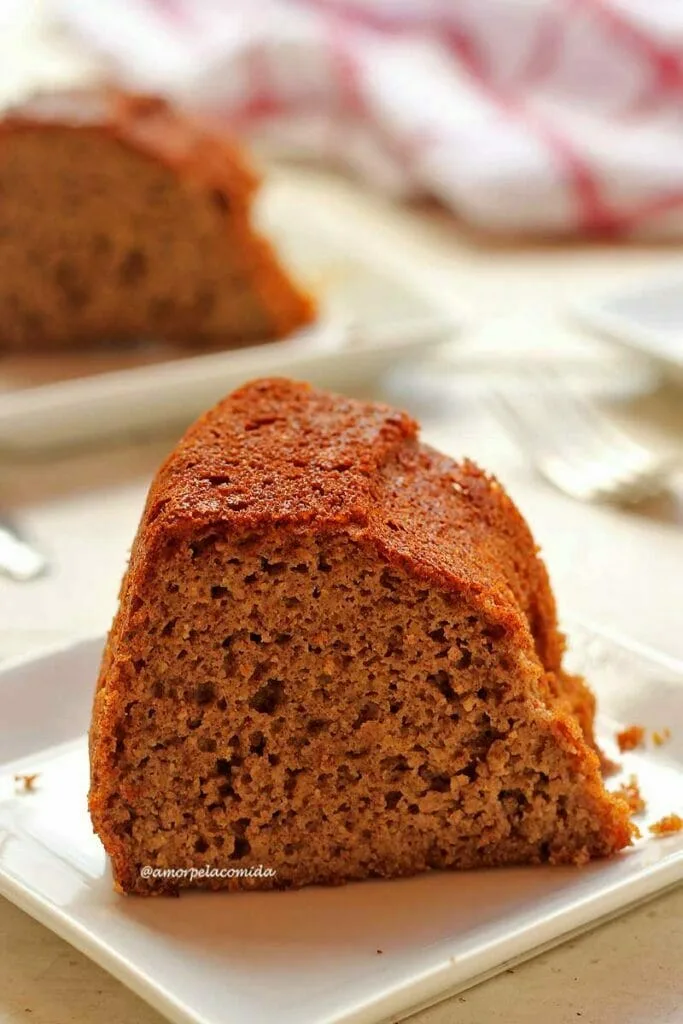 Un morceau de gâteau sur une petite assiette carrée sur une table blanche, en arrière-plan une partie d'une autre tranche de gâteau apparaît sur l'assiette