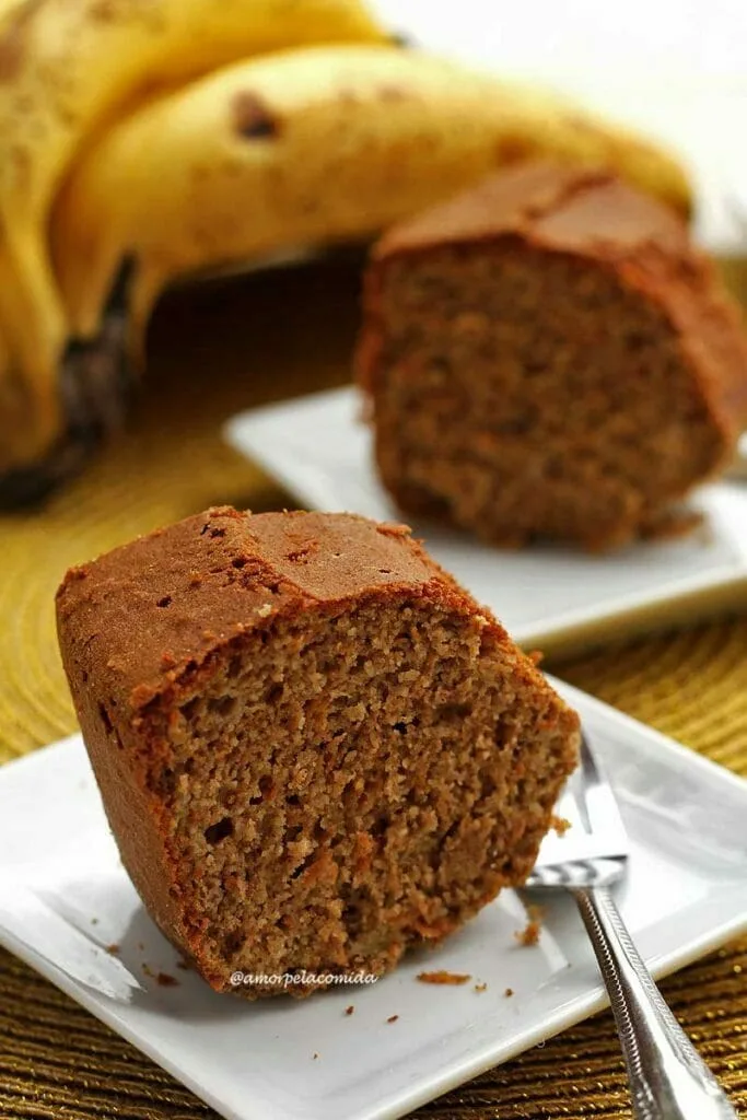 Deux tranches de gâteau à la banane sur une petite assiette carrée, à côté de chaque assiette une petite fourchette en métal et en arrière-plan un régime de banane