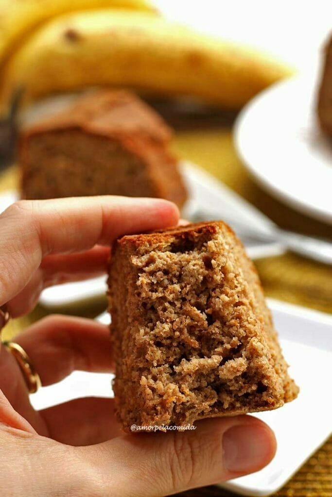 Mão segurando pedaço de bolo de banana mordido mostrando a textura macia