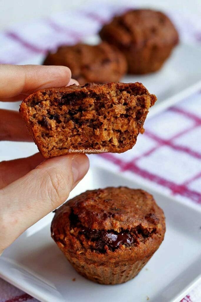 Mão segurando bolinho pequeno que está mordido, a textura é macia e tem alguns pedacinhos de chocolate, abaixo um bolinho pequeno inteiro