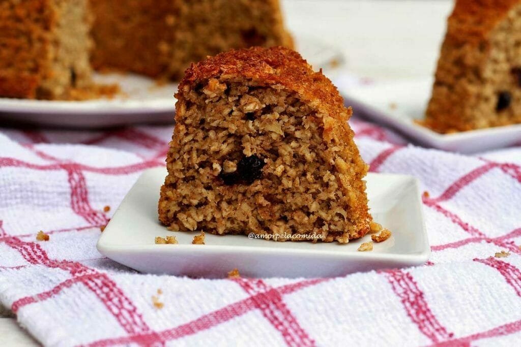 Pedaço de bolo de maçã sobre prato quadrado pequeno, ao fundo o bolo partido