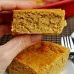 Mão segurando pedaço quadrado de bolo de fubá, abaixo tem mais um pedaço sobre um prato branco pequeno