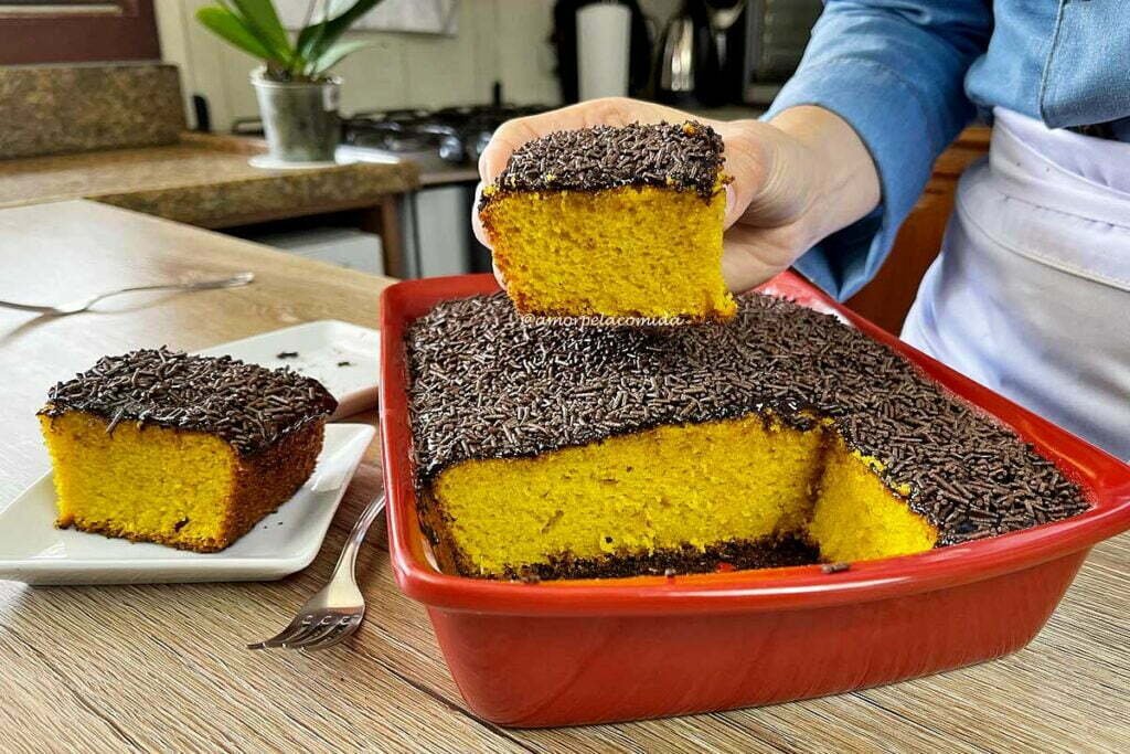 Forma de bolo retangular com bolo de cenoura com cobertura de chocolate dentro, ao lado um pedaço cortado e acima da forma uma mão segurando um pedaço de bolo