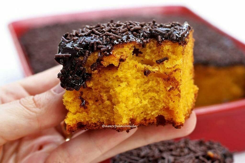 Mão segurando pedaço quadrado de bolo de cenoura com cobertura de chocolate que foi mordido