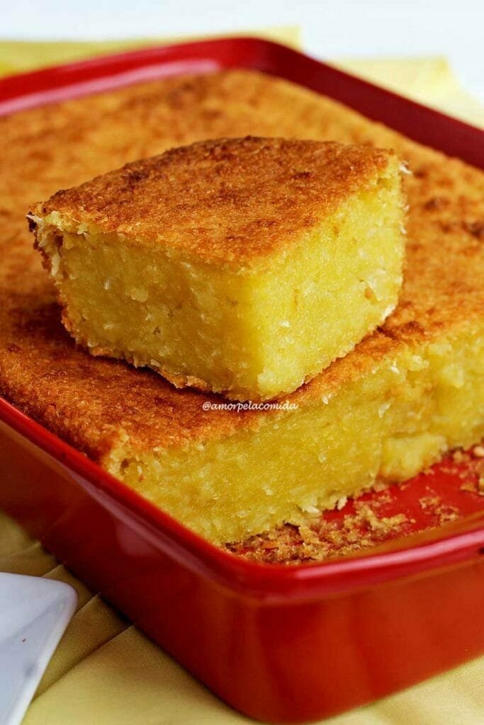 Pedaço de bolo quadrado de mandioca cremoso que está apoiado sobre o bolo de mandioca que está dentro da forma