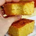 Mão segurando pedaço de bolo de mandioca cremoso que já foi mordido, abaixo um pedaço de bolo sobre um prato quadrado pequeno