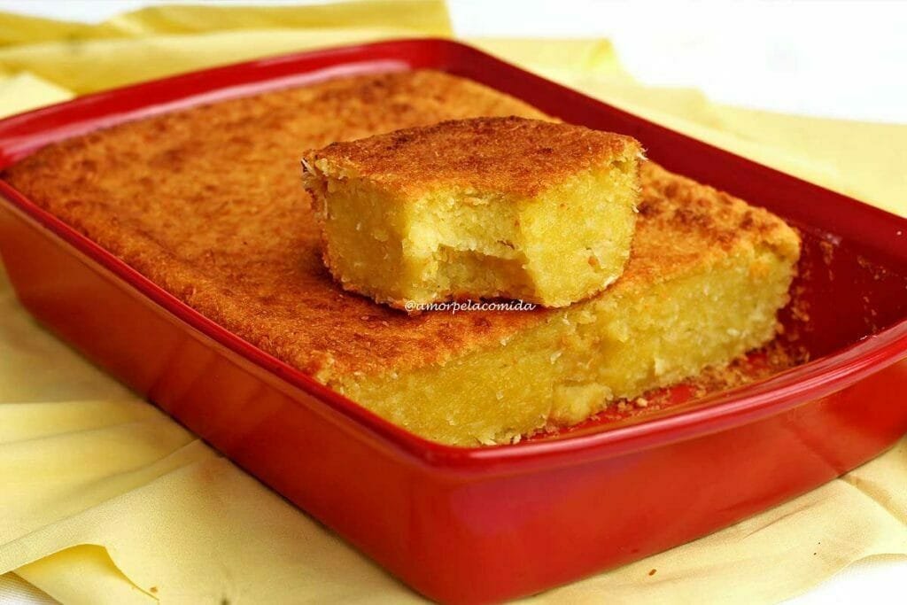 Pedaço de bolo quadrado de mandioca cremoso que está apoiado sobre o bolo de mandioca que está dentro da forma