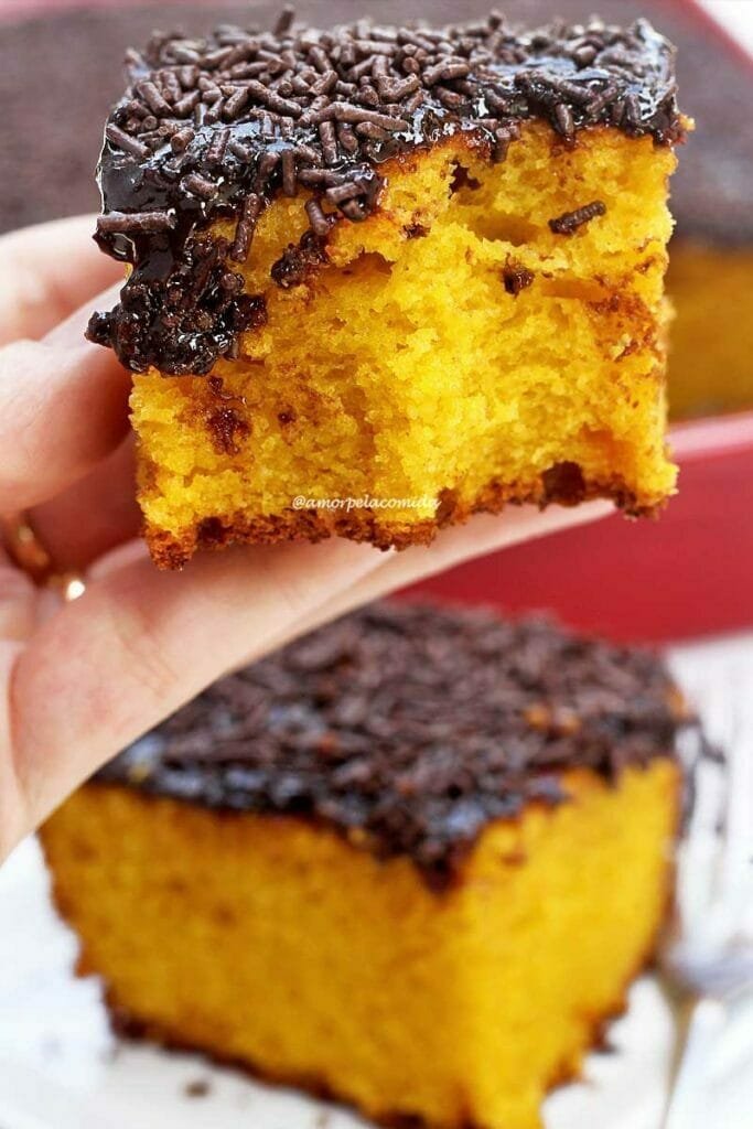 Mão segurando pedaço de bolo de cenoura com cobertura de chocolate que já foi mordido, abaixo mais um pedaço sobre prato quadrado pequeno