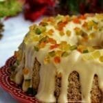 Bolo redondo com furo no meio sobre prato da cor vinho, o bolo tem uma cobertura cremosa e está decorados com frutas cristalizadas, ao fundo enfeites natalinos