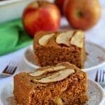 Dois pedaços retangulares de bolo de maçã com aveia, no topo fatias finas de maçã enfeitando o pedaço, ao fundo maçãs inteiras sobre mesa branca