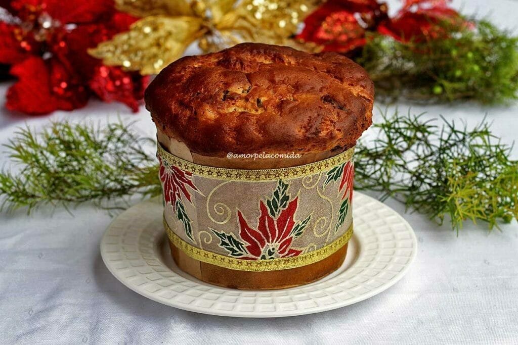 Panetone sobre prato redondo branco, ao redor do panetone uma fita com flores natalinas decorando a receita, atrás enfeites de natal