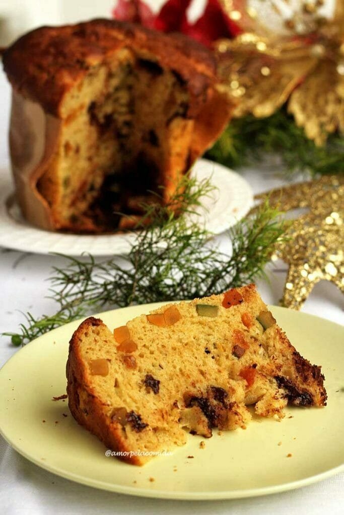 Fatia de panetone de frutas com gotas de chocolate sobre prato redondo verde-claro, ao fundo o panetone cortado e enfeites de natal