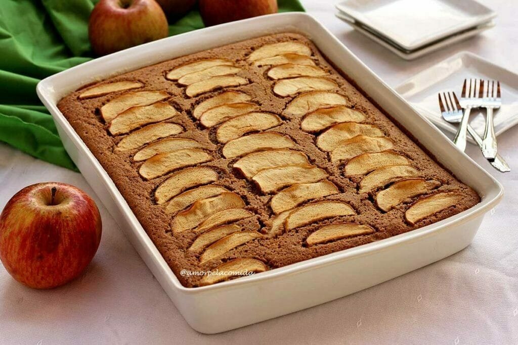 Bolo de maçã com fatias de maçã decorando o topo dentro de uma forma de cerâmica retangular da cor branca, algumas maçã ao redor e pratinhos quadrados ao lado